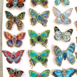 Puede incluir: Una colecci&oacute;n de 18 dijes de mariposa hechos a mano con alas iridiscentes y detalles de joyas. Los dijes est&aacute;n dispuestos en filas sobre una superficie blanca.