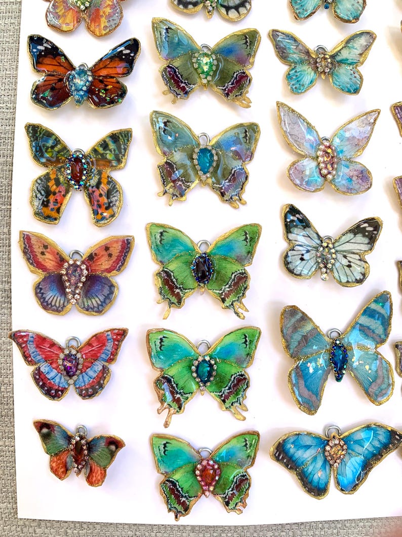 Puede incluir: Una colecci&oacute;n de 18 dijes de mariposa hechos a mano con alas iridiscentes y detalles de joyas. Los dijes est&aacute;n dispuestos en filas sobre una superficie blanca.
