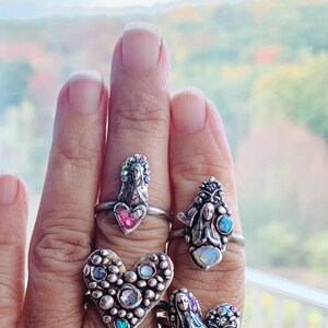 May include: Four silver rings with religious motifs are shown on a hand. Each ring features a saint or angel figure, embellished with colourful gemstones and set within a heart or oval shape. The rings are set against a blurred background.