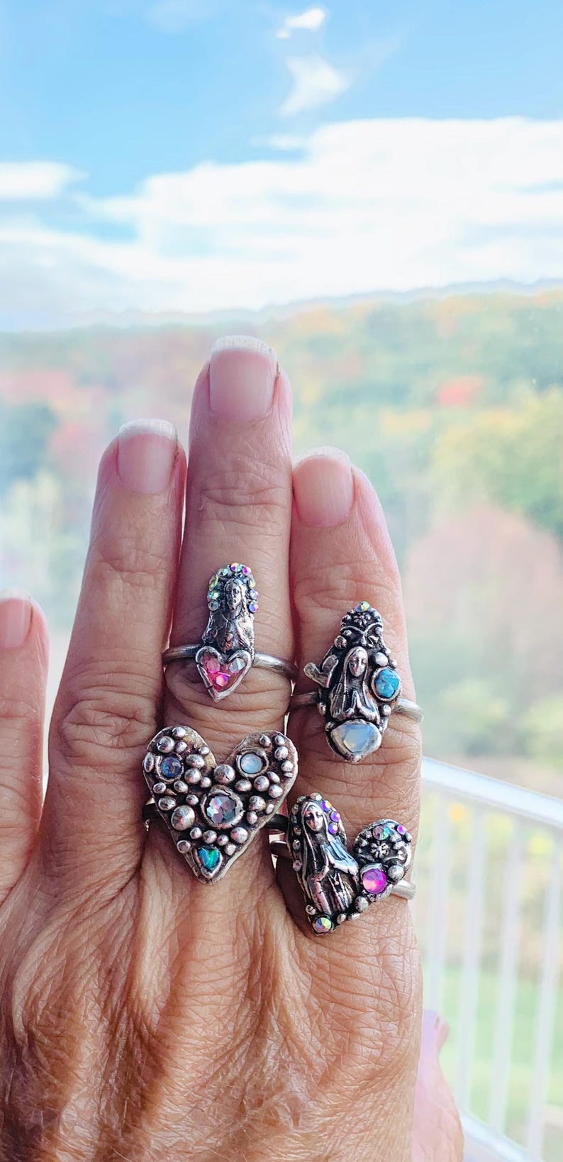 May include: Four silver rings with religious motifs are shown on a hand. Each ring features a saint or angel figure, embellished with colourful gemstones and set within a heart or oval shape. The rings are set against a blurred background.