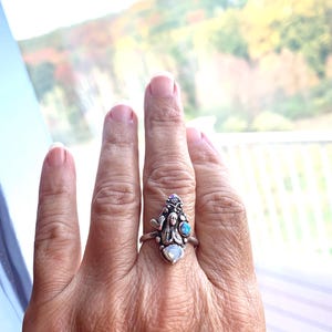 May include: A silver ring with an intricate design, including a central female figure and small gemstones. The ring incorporates a pearlescent stone and a blue gemstone. The background is a blurred outdoor scene, with autumn colours.