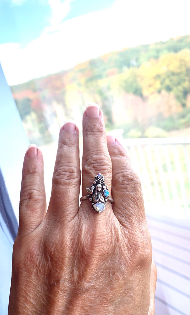 May include: A silver ring with an intricate design, including a central female figure and small gemstones. The ring incorporates a pearlescent stone and a blue gemstone. The background is a blurred outdoor scene, with autumn colours.
