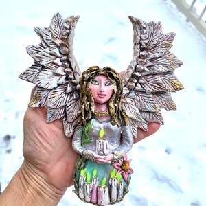 May include: A handmade ceramic angel figurine with wings, holding a candle. The angel is wearing a green dress and has a flower on her dress. The angel is painted in shades of pink, white, and gold.