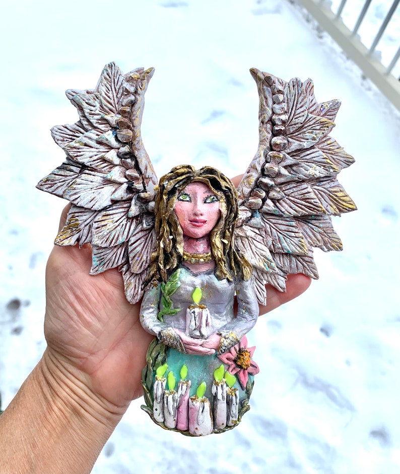 May include: A handmade ceramic angel figurine with wings, holding a candle. The angel is wearing a green dress and has a flower on her dress. The angel is painted in shades of pink, white, and gold.