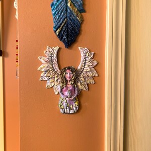 May include: A ceramic angel figurine with wings made of leaves, holding a candle. The angel is painted in shades of pink, purple, and gold. The figurine is mounted on a light orange wall.