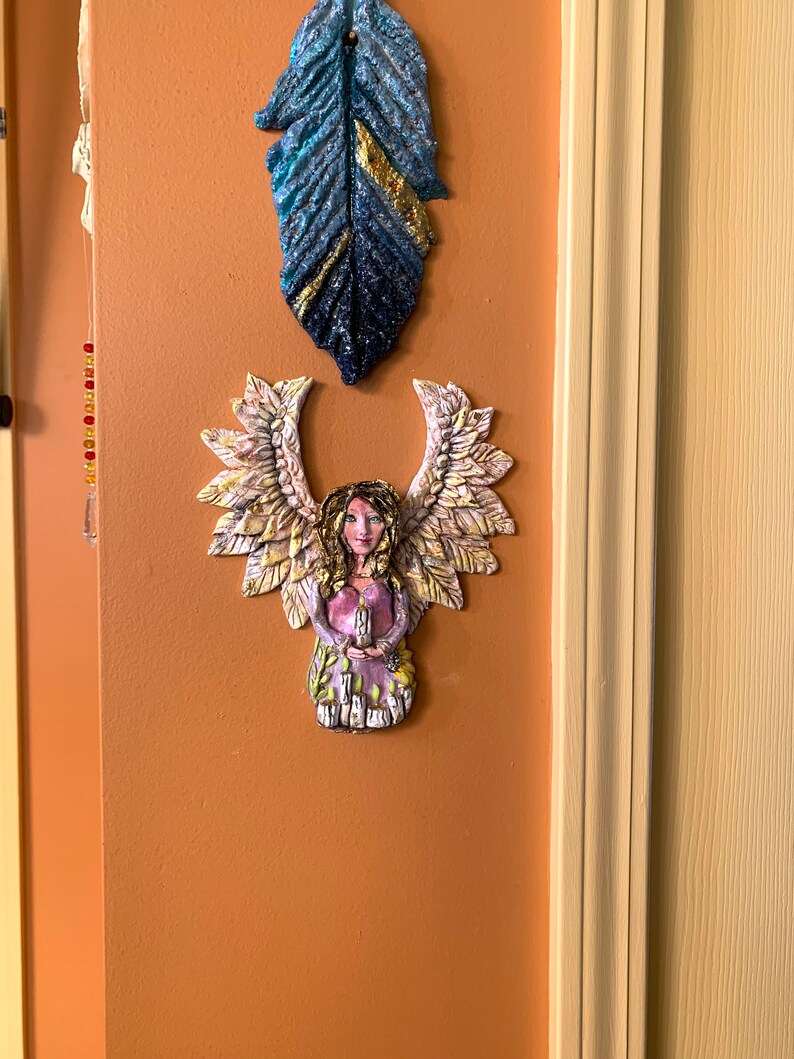May include: A ceramic angel figurine with wings made of leaves, holding a candle. The angel is painted in shades of pink, purple, and gold. The figurine is mounted on a light orange wall.