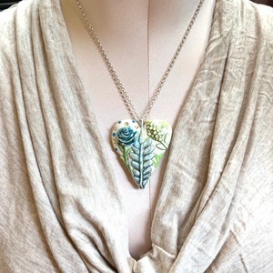 Puede incluir: Un collar de cadena plateada con un colgante blanco en forma de coraz&oacute;n. El colgante est&aacute; decorado con un dise&ntilde;o floral azul y verde.