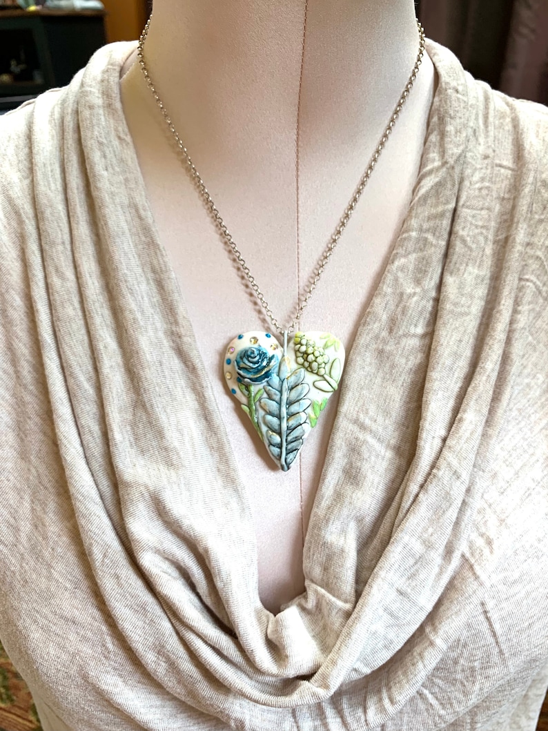 Puede incluir: Un collar de cadena plateada con un colgante blanco en forma de coraz&oacute;n. El colgante est&aacute; decorado con un dise&ntilde;o floral azul y verde.