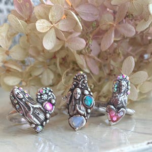 May include: Three silver rings with intricate designs and iridescent gemstones. The rings feature heart, figure, and face motifs. The background includes dried flowers with a soft pink and cream colour palette, creating a vintage aesthetic.