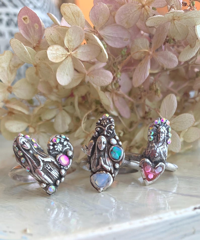 May include: Three silver rings with intricate designs and iridescent gemstones. The rings feature heart, figure, and face motifs. The background includes dried flowers with a soft pink and cream colour palette, creating a vintage aesthetic.