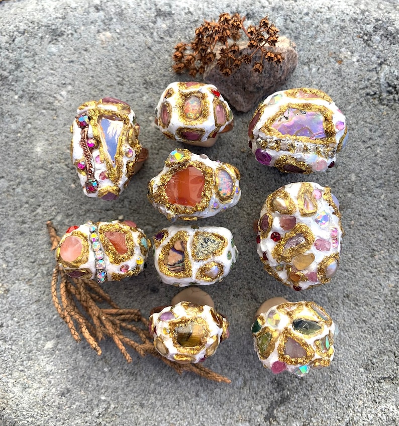 May include: Nine white rings with gold accents and various colorful stones and crystals. The rings are arranged in a circle on a gray textured surface.