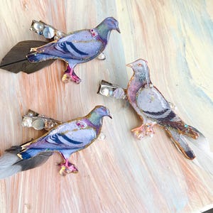 May include: Three hair clips featuring pigeon-shaped ornaments with iridescent purple, blue, and grey plumage. Each bird is accented with gold glitter, small white pearls, and attached natural feathers in shades of grey, brown, and white.