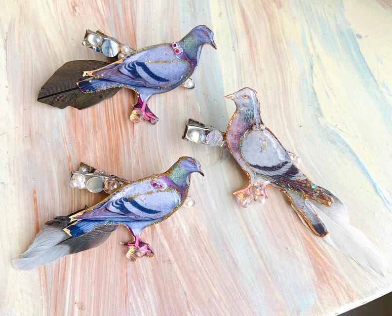 May include: Three hair clips featuring pigeon-shaped ornaments with iridescent purple, blue, and grey plumage. Each bird is accented with gold glitter, small white pearls, and attached natural feathers in shades of grey, brown, and white.