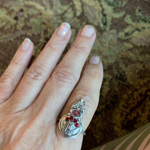 K&ouml;nnte beinhalten: Ein silberner Ring mit einem roten Stein und einem detaillierten Design. Der Ring wird an einem Finger mit klarem Nagellack getragen.
