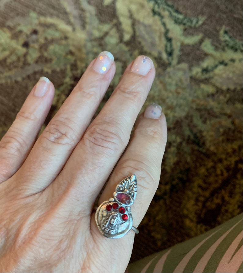 K&ouml;nnte beinhalten: Ein silberner Ring mit einem roten Stein und einem detaillierten Design. Der Ring wird an einem Finger mit klarem Nagellack getragen.