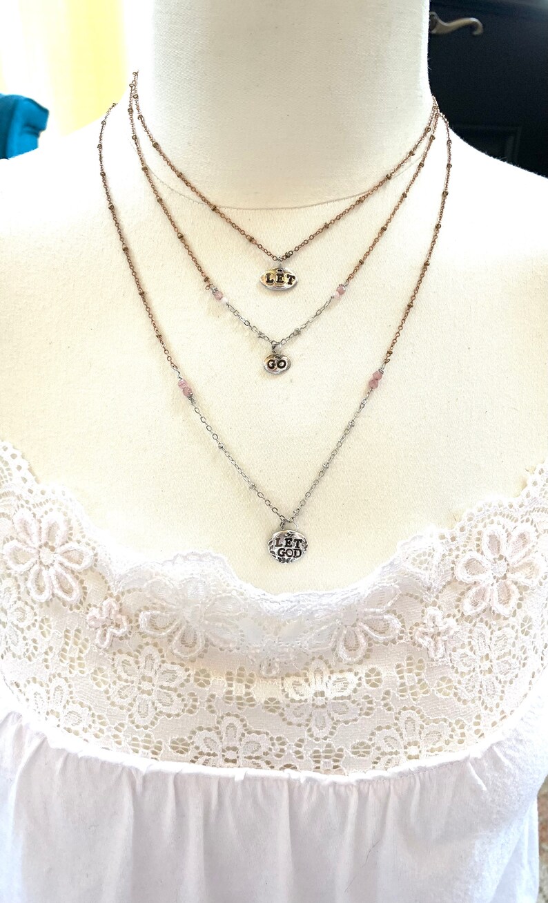 Puede incluir: Collar de capas con tres dijes de plata. Los dijes dicen "Let", "Go" y "Let God".