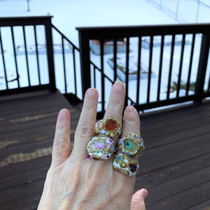 May include: Four colorful rings with gold accents and various gemstones. The rings are stacked on a person's finger.