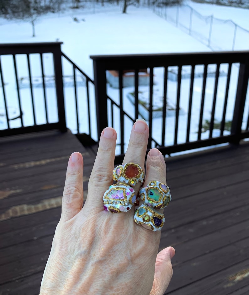 May include: Four colorful rings with gold accents and various gemstones. The rings are stacked on a person's finger.