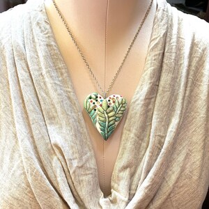 Puede incluir: Un collar de cadena de plata con un colgante de coraz&oacute;n de cer&aacute;mica verde, blanco y azul. El coraz&oacute;n tiene un dise&ntilde;o detallado de hojas y flores con acentos coloridos.