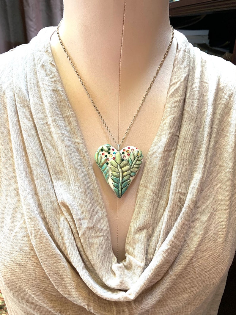 Puede incluir: Un collar de cadena de plata con un colgante de coraz&oacute;n de cer&aacute;mica verde, blanco y azul. El coraz&oacute;n tiene un dise&ntilde;o detallado de hojas y flores con acentos coloridos.