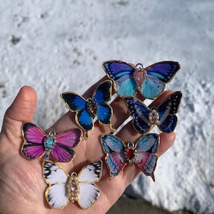 Puede incluir: Seis abalorios de mariposa de resina con detalles dorados y adornos de piedras preciosas. Las mariposas son de varios colores, incluyendo azul, morado, rosa y blanco.