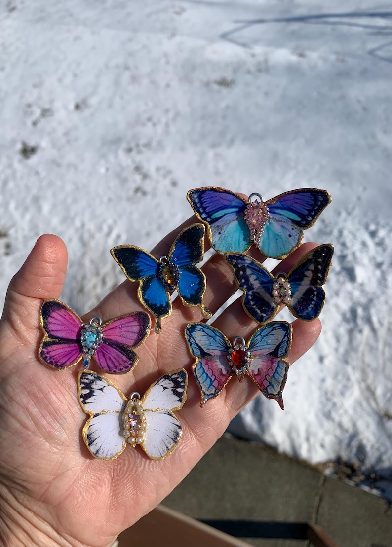 Puede incluir: Seis abalorios de mariposa de resina con detalles dorados y adornos de piedras preciosas. Las mariposas son de varios colores, incluyendo azul, morado, rosa y blanco.