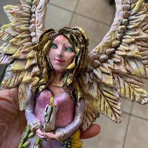 May include: A handmade angel figurine with wings made of textured paper mache. The angel is holding a candle and has a pink dress with gold accents. The angel is standing on a base with candles and sunflowers.