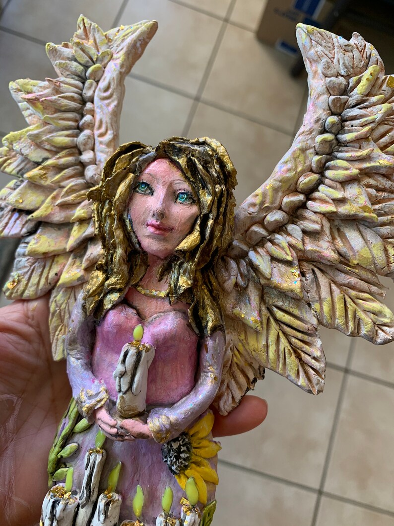 May include: A handmade angel figurine with wings made of textured paper mache. The angel is holding a candle and has a pink dress with gold accents. The angel is standing on a base with candles and sunflowers.