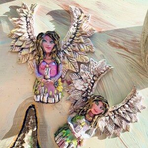 May include: Two ceramic angel figurines with wings made of leaves. The angels are holding candles and have painted faces with green eyes. The angels are sitting on a light brown surface.