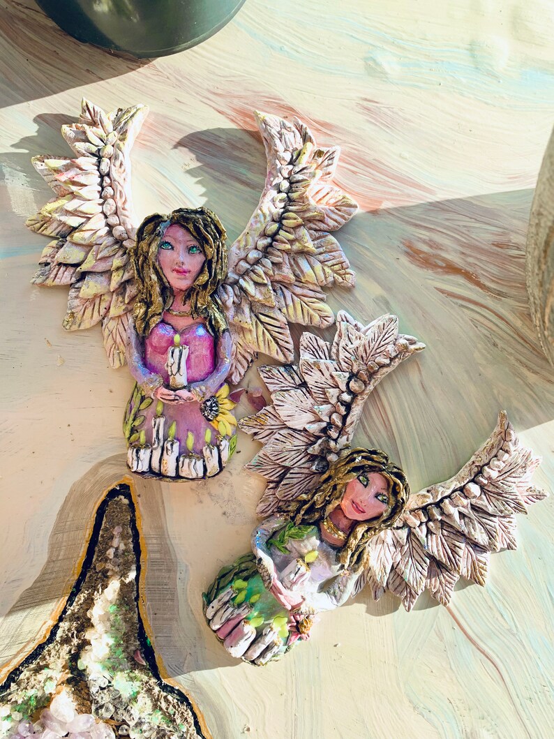 May include: Two ceramic angel figurines with wings made of leaves. The angels are holding candles and have painted faces with green eyes. The angels are sitting on a light brown surface.
