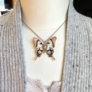 Puede incluir: Un collar de cadena dorada con un colgante de mariposa. La mariposa est&aacute; hecha de porcelana blanca con detalles negros y morados y tiene un centro dorado.