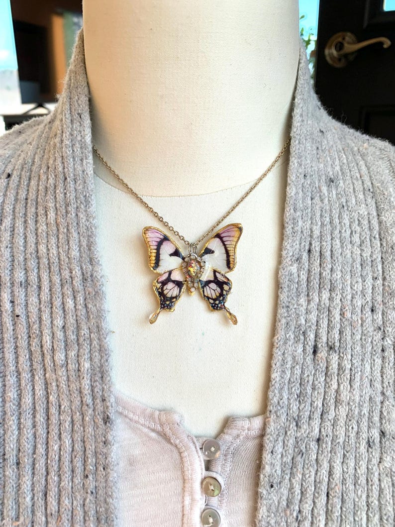 Puede incluir: Un collar de cadena dorada con un colgante de mariposa. La mariposa est&aacute; hecha de porcelana blanca con detalles negros y morados y tiene un centro dorado.
