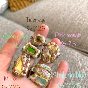 May include: Five handmade rings with different stone and glass inlays. The rings are made of white clay with gold accents. The stones include tiger eye, green glass, pink mosaic, mirror, and abalone blue. The rings are sized 7.25 and 7.75.