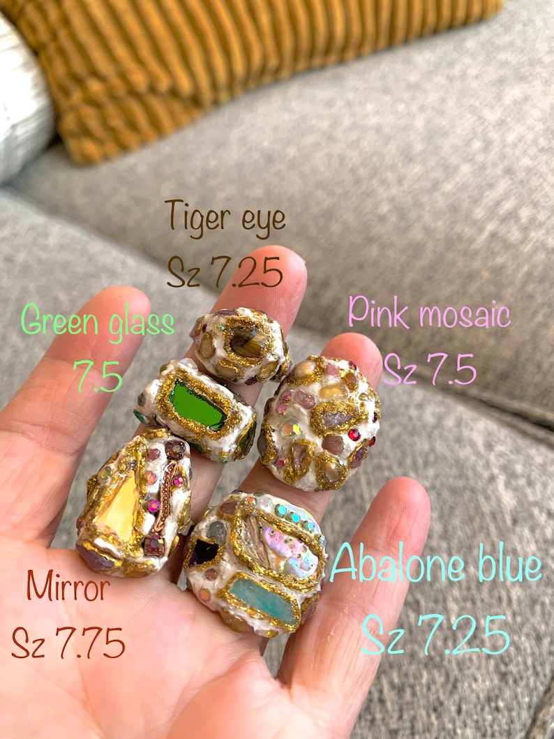 May include: Five handmade rings with different stone and glass inlays. The rings are made of white clay with gold accents. The stones include tiger eye, green glass, pink mosaic, mirror, and abalone blue. The rings are sized 7.25 and 7.75.