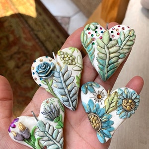 Puede incluir: Cuatro adornos de coraz&oacute;n de cer&aacute;mica hechos a mano con dise&ntilde;os florales y detalles dorados. Cada coraz&oacute;n presenta diferentes flores y hojas, incluyendo una rosa azul, flores amarillas y hojas verdes. Los corazones est&aacute;n decorados con diamantes de imitaci&oacute;n y tienen un fondo blanco.