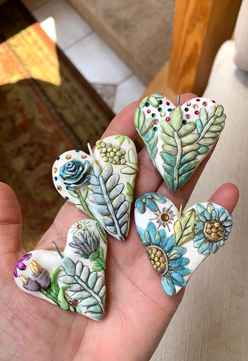 Puede incluir: Cuatro adornos de coraz&oacute;n de cer&aacute;mica hechos a mano con dise&ntilde;os florales y detalles dorados. Cada coraz&oacute;n presenta diferentes flores y hojas, incluyendo una rosa azul, flores amarillas y hojas verdes. Los corazones est&aacute;n decorados con diamantes de imitaci&oacute;n y tienen un fondo blanco.