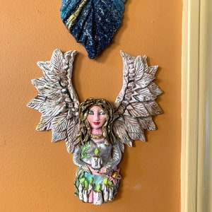 May include: A ceramic angel figurine with wings, holding a candle. The angel has a white dress with green accents and a floral base. The figurine is painted with gold accents and has a realistic face.