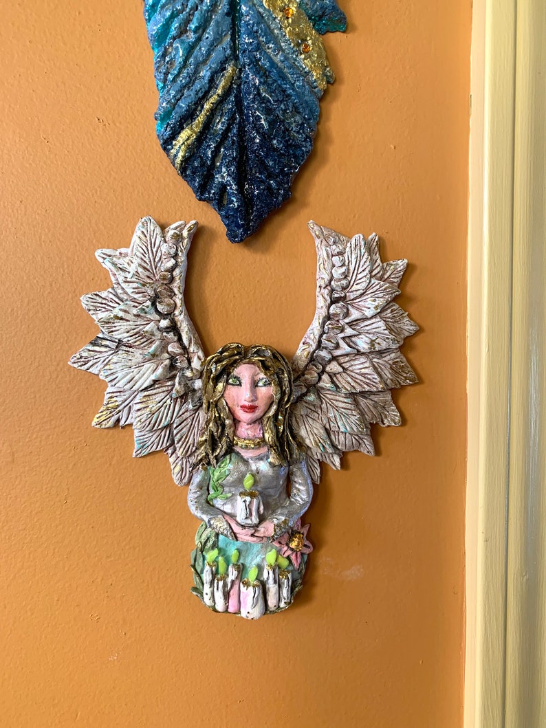 May include: A ceramic angel figurine with wings, holding a candle. The angel has a white dress with green accents and a floral base. The figurine is painted with gold accents and has a realistic face.