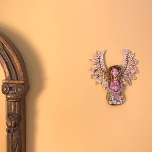 May include: A ceramic angel figurine with wings made of leaves. The angel is wearing a purple dress and has a crown of flowers on her head.