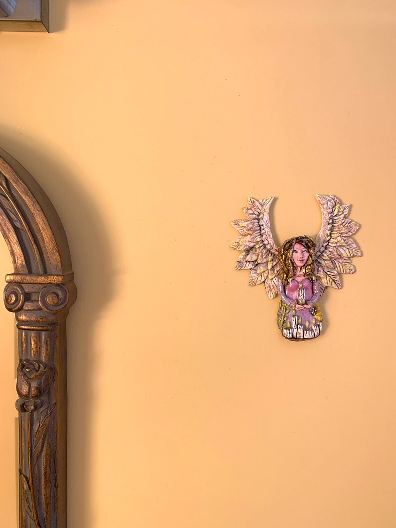 May include: A ceramic angel figurine with wings made of leaves. The angel is wearing a purple dress and has a crown of flowers on her head.