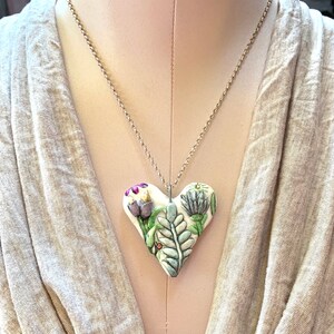 Puede incluir: Un collar con un colgante de coraz&oacute;n de cer&aacute;mica blanca y una cadena de plata. El coraz&oacute;n est&aacute; decorado con un dise&ntilde;o floral con hojas verdes, flores moradas y flores blancas.