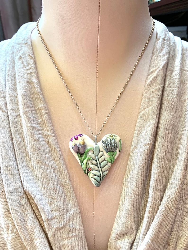 Puede incluir: Un collar con un colgante de coraz&oacute;n de cer&aacute;mica blanca y una cadena de plata. El coraz&oacute;n est&aacute; decorado con un dise&ntilde;o floral con hojas verdes, flores moradas y flores blancas.