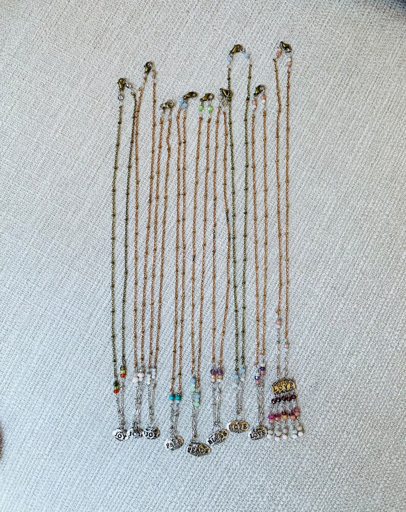 Puede incluir: Una colecci&oacute;n de 12 collares de cadena dorada con cuentas y dijes de varios colores. Cada dije tiene una palabra diferente, incluyendo "Alegr&iacute;a", "Paz", "Amor" y "Esperanza".