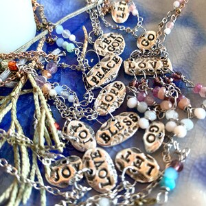 Puede incluir: Una colecci&oacute;n de collares con dijes de plata con varias palabras en los dijes, incluyendo "Let God", "Peace", "Hope", "Bless", "Love", "Joy", "Let Go", y "Go". Los collares est&aacute;n sobre un fondo azul con peque&ntilde;as cuentas de colores.