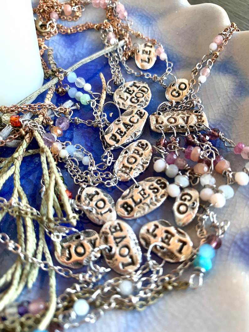 Puede incluir: Una colecci&oacute;n de collares con dijes de plata con varias palabras en los dijes, incluyendo "Let God", "Peace", "Hope", "Bless", "Love", "Joy", "Let Go", y "Go". Los collares est&aacute;n sobre un fondo azul con peque&ntilde;as cuentas de colores.