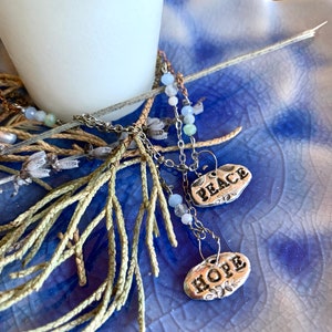 Puede incluir: Un collar de cadena de plata con dos dijes de plata que dicen "Peace" y "Hope". El collar est&aacute; sobre una superficie con estampado azul y blanco con una rama de follaje verde.