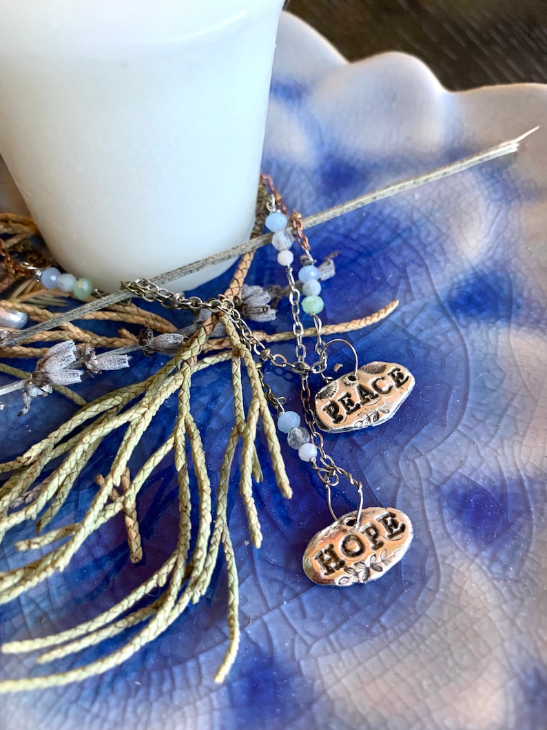 Puede incluir: Un collar de cadena de plata con dos dijes de plata que dicen "Peace" y "Hope". El collar est&aacute; sobre una superficie con estampado azul y blanco con una rama de follaje verde.