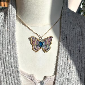 Puede incluir: Un collar de cadena dorada con un colgante de mariposa. La mariposa est&aacute; hecha de papel y tiene una gema azul en el centro. La mariposa es de color rosa, morado y amarillo.