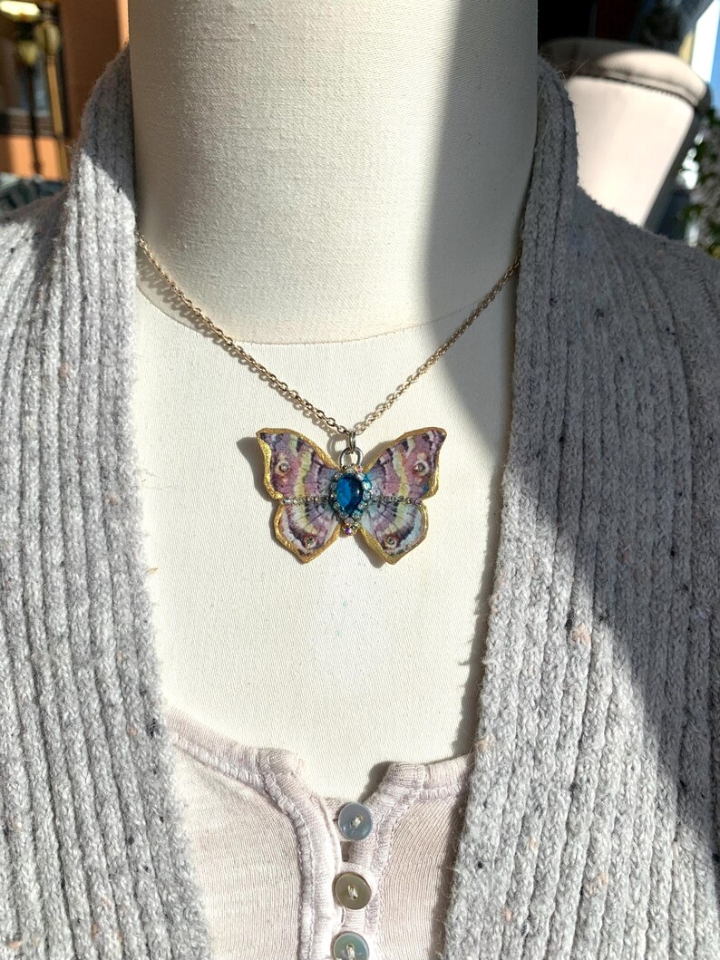 Puede incluir: Un collar de cadena dorada con un colgante de mariposa. La mariposa est&aacute; hecha de papel y tiene una gema azul en el centro. La mariposa es de color rosa, morado y amarillo.