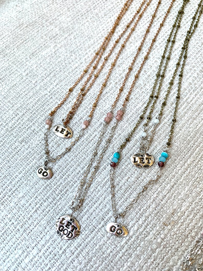 Puede incluir: Cuatro collares de plata con diferentes dijes. Los dijes dicen "Let Go", "Let God" y "Let" con un peque&ntilde;o dije de cuentas. Los collares est&aacute;n sobre un fondo de tela blanca.